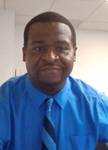 A photo of Ray McCoy, a Black man with short hair and a mustache. Ray is smiling and wearing a bright blue shirt and striped blue tie.