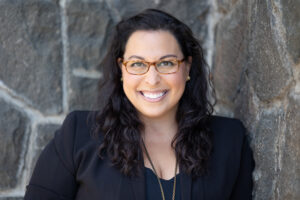 Headshot of Melissa Doman, a light-skinned woman with long dark hair and glasses. Melissa is standing in front of a brick wall, wearing a black blazer, and smiling at the camera.