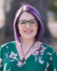 Headshot of Dr. Allison Battles, a white woman with shoulder-length purple hair. Allison is wearing glasses, a necklace, and a green floral blouse.