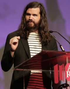 Photo of Kevin Rumley, a white man with long brown hair and a beard. Kevin is standing at a podium, wearing a striped white shirt and a black blazer. 