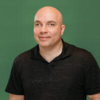 Headshot of Russell Shaffer, a white man with a shaved head. Russell is wearing a black collared t-shirt and is smiling at the camera.