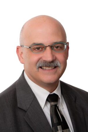 Headshot of Sam Catanzaro. Sam has grey glasses and a grey mustache. Sam is wearing a dark suite and tie and is smiling at the camera.