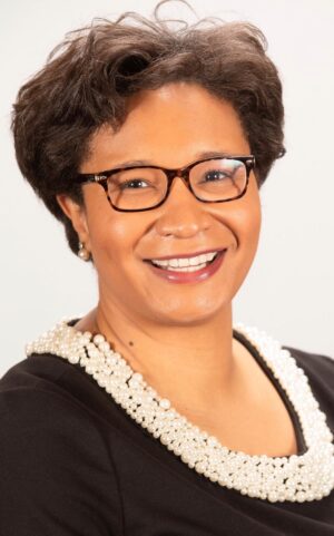 Headshot of J'Nai Buchanan. J'Nai has short brown hair and glasses. She is wearing a black top with white pearl collar and is smiling at the camera.