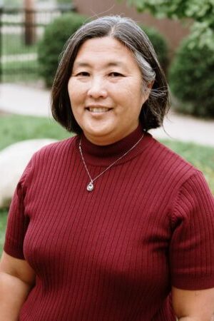 Headshot of Kari Scanlon. Kari has short dark hair and is standing outdoors in a garden area. Kari is wearing a red turtleneck and is smiling at the camera.