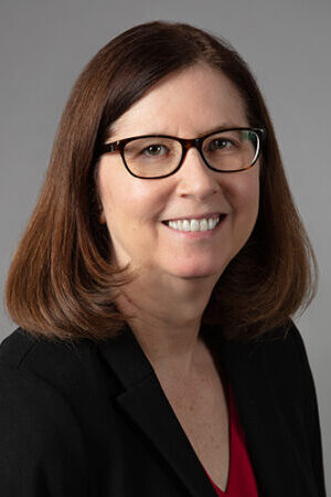 Headshot of Lisa Stock. Lisa has shoulder length brown hair, glasses, and is wearing a black blazer. She is smiling at the camera.