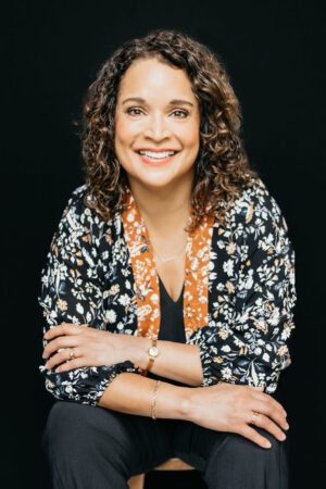 Headshot of Maria McFarlane. Maria has short brown curly hair and is wearing a black and orange floral print blazer. Maria is smiling at the camera.